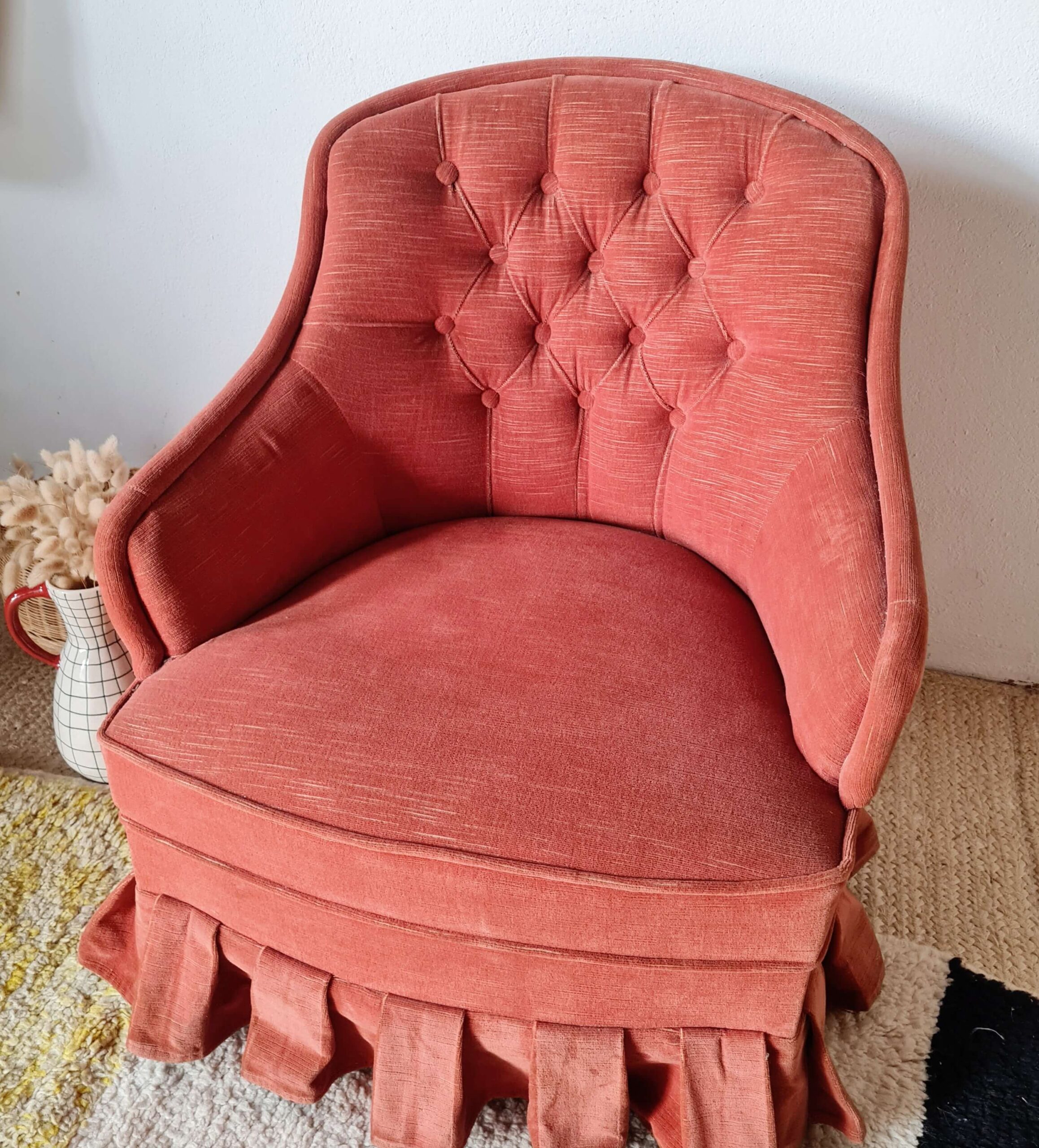 Vue de trois quarts d'un fauteuil vintage capitonné en tissu rouge terracotta avec une jupe plissée.