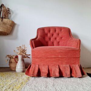 Fauteuil crapaud vintage capitonné en tissu rouge terracotta, avec jupe plissée à volants, installé dans un salon à la décoration bohème.