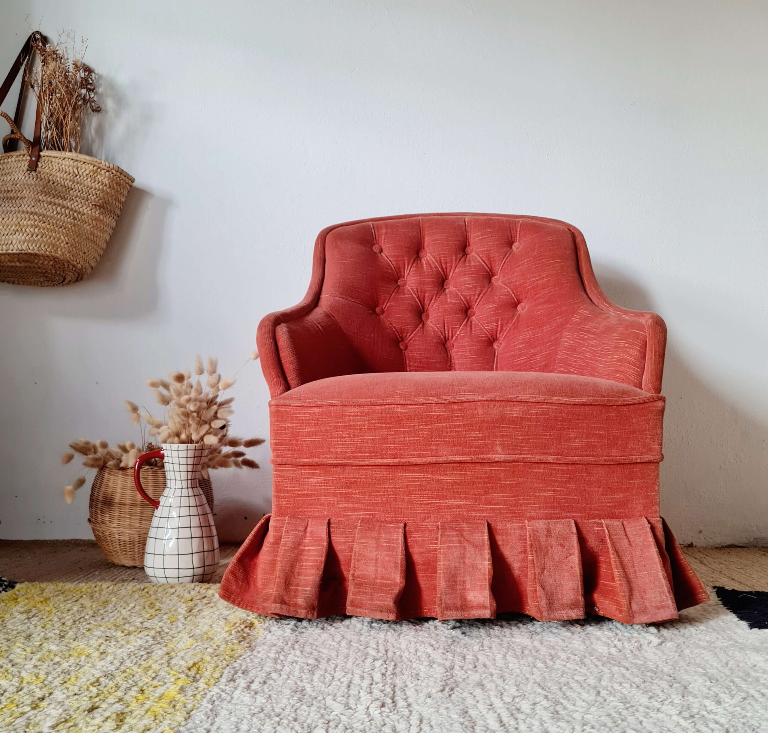 Fauteuil crapaud vintage capitonné en tissu rouge terracotta, avec jupe plissée à volants, installé dans un salon à la décoration bohème.