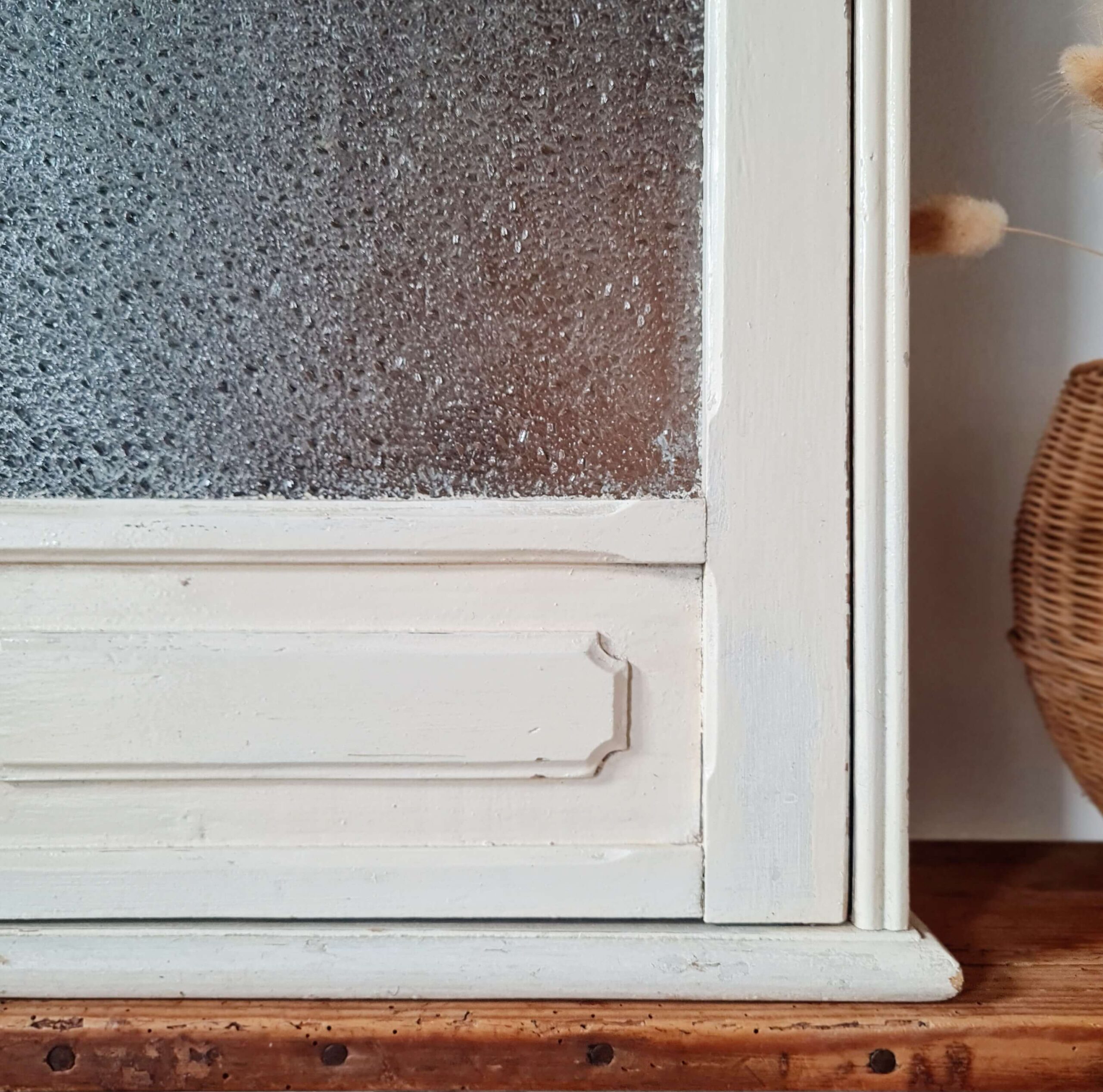 Gros plan sur le coin inférieur droit de la porte vitrée et du cadre de l'armoire murale, soulignant la texture du verre et l'usure naturelle du bois peint.