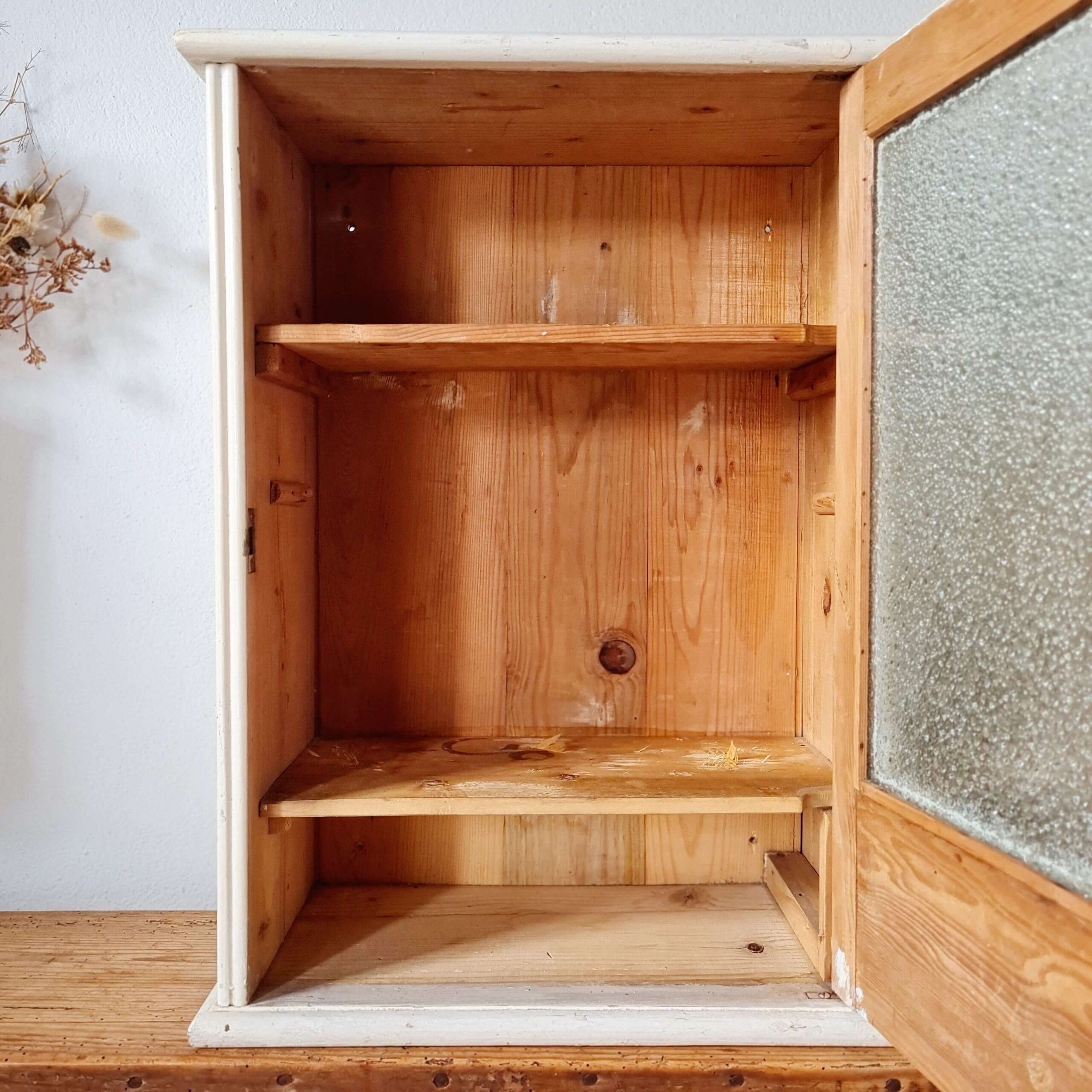 La porte de l'armoirette ancienne est ouverte, révélant l'intérieur en bois brut chaleureux doté de deux étagères et d'un fond en planches verticales.