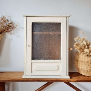Vue de face de la petite armoire murale vitrée ancienne, encadrée par un décor épuré avec un panier en osier rempli de fleurs séchées sur la droite.