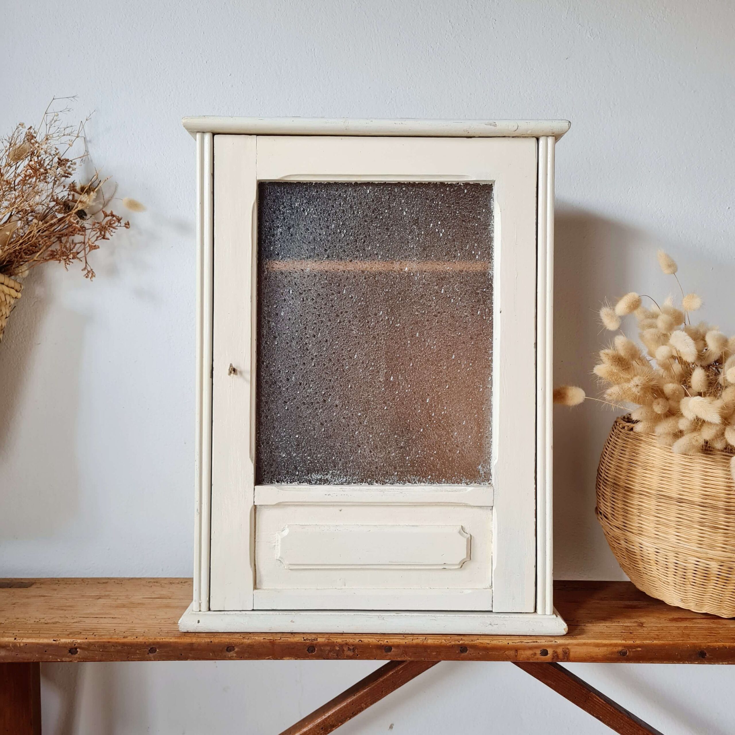 Vue de face de la petite armoire murale vitrée ancienne, encadrée par un décor épuré avec un panier en osier rempli de fleurs séchées sur la droite.