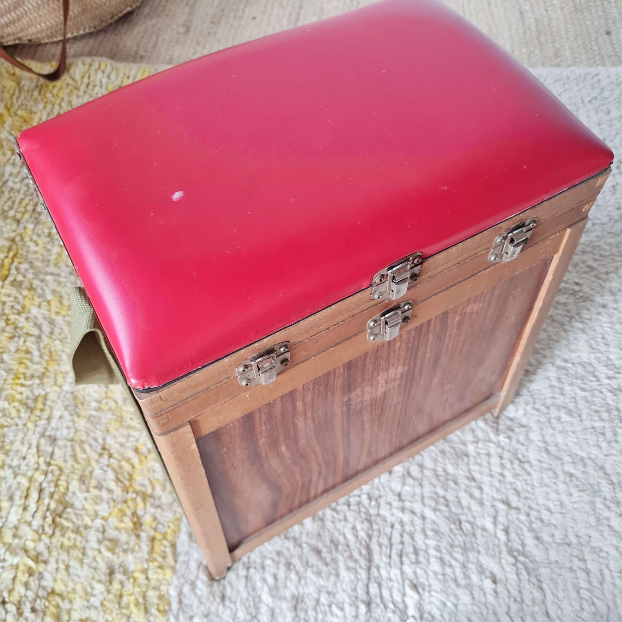 Autre prise de vue de l'ancien tabouret de pêcheur, vue de trois quarts depuis le haut. Elle montre l'ensemble du meuble, son coffre et son assise rouge sur le tapis.