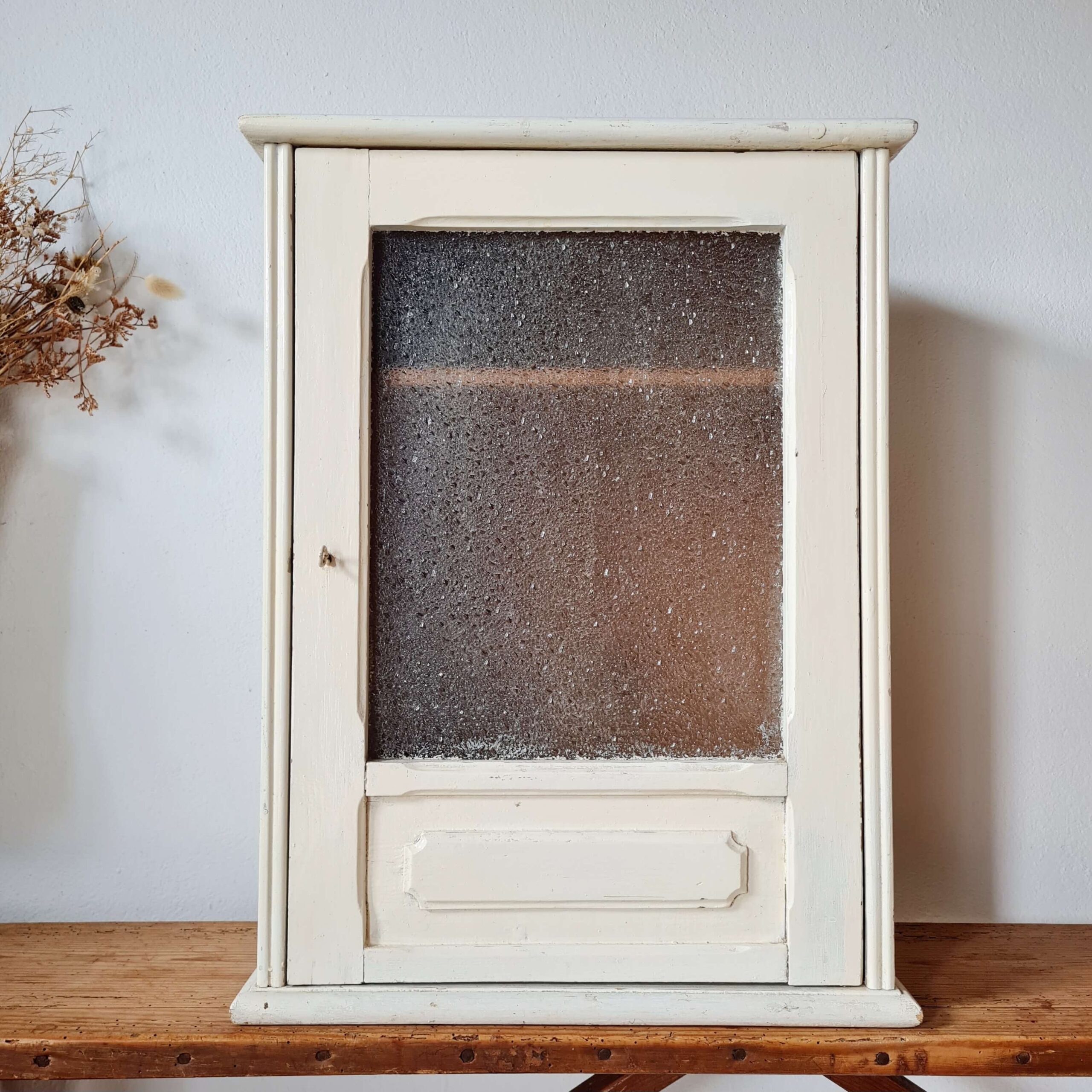 Petite vitrine murale ancienne repeinte en blanc cassé, posée sur un meuble en bois, avec une porte vitrée texturée et un panneau bas mouluré.