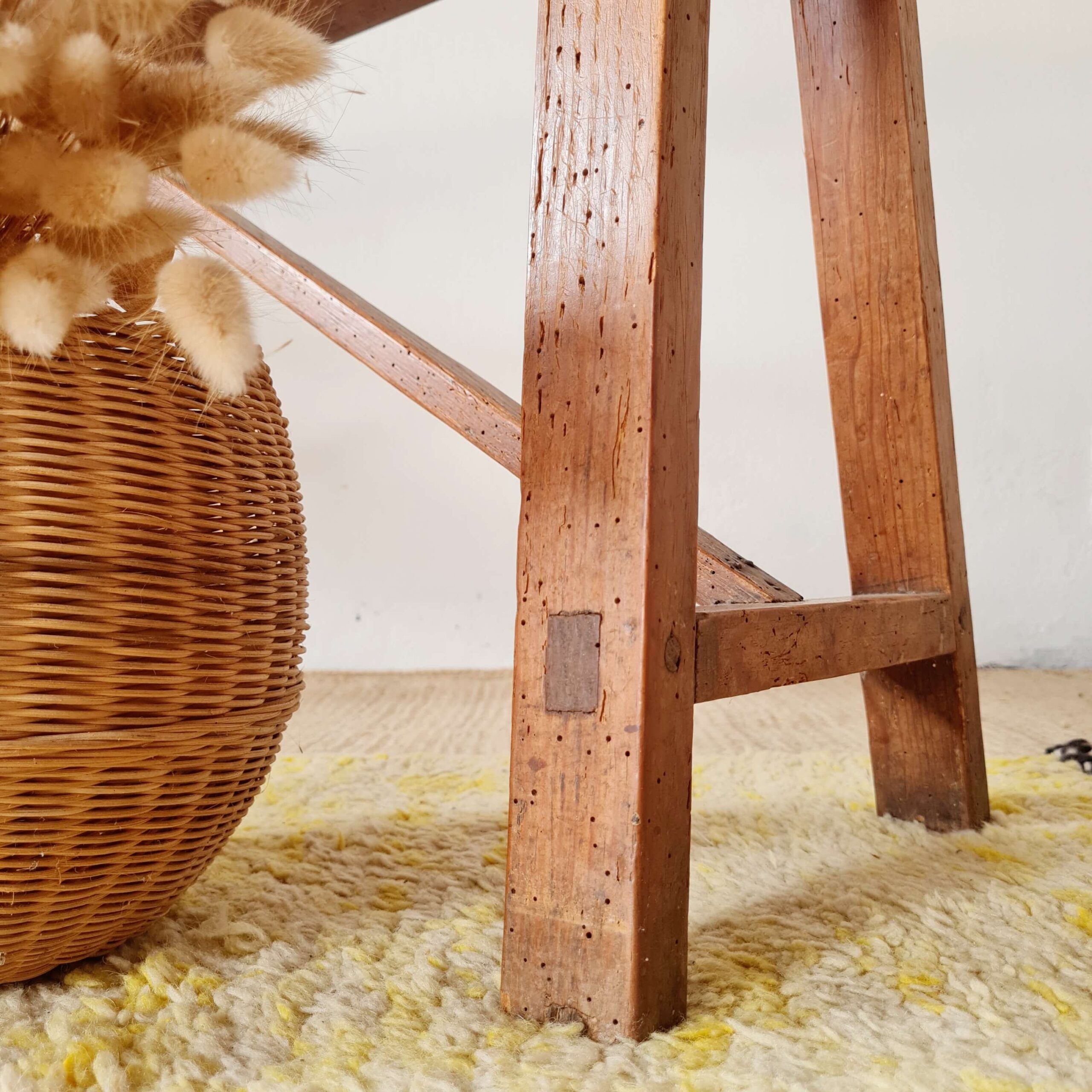 Détail du piètement rustique en bois massif d'un banc ancien, à côté d'un grand vase en vannerie tressée posé sur un tapis en laine
