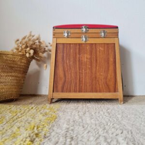 Ancien tabouret de pêcheur en bois avec un coffre de rangement intégré et une assise en simili cuir rouge. Le meuble est fermé par des fermoirs métalliques et posé sur un tapis berbère.
